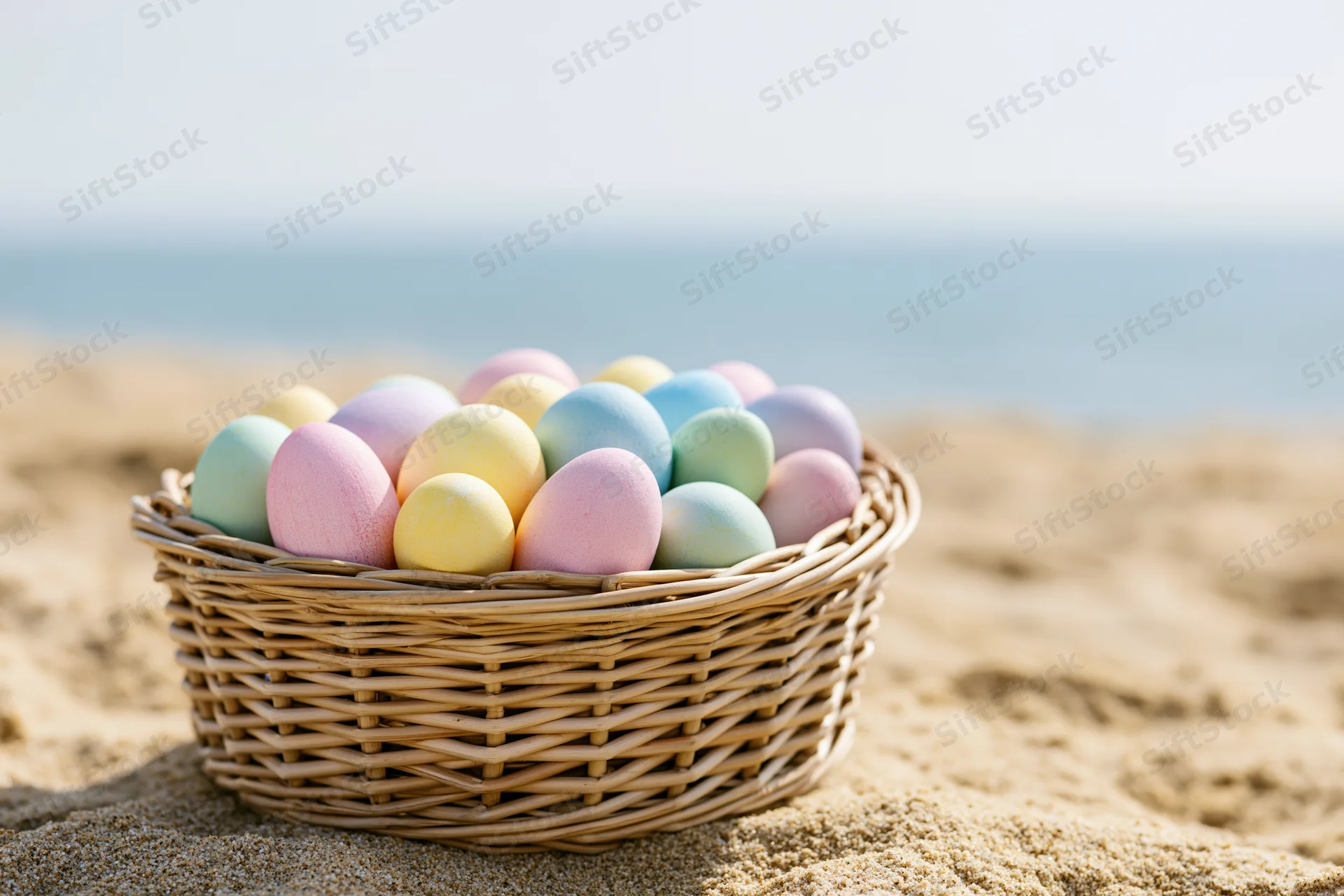 Wicker basket filled with pastel-colored easter eggs on sandy beach near ocean