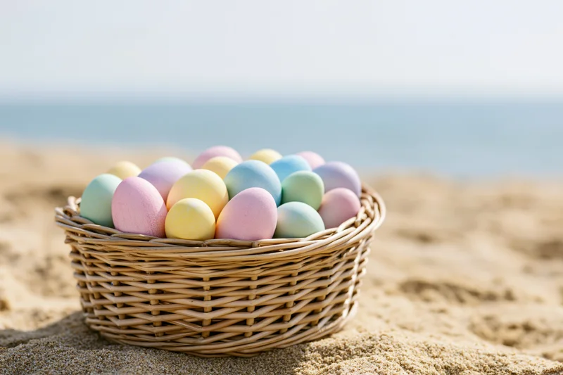 Wicker basket filled with pastel-colored easter eggs on sandy beach near ocean