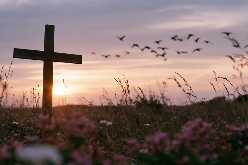Wooden christian cross standing in a wildflower meadow at sunset with birds flying overhead