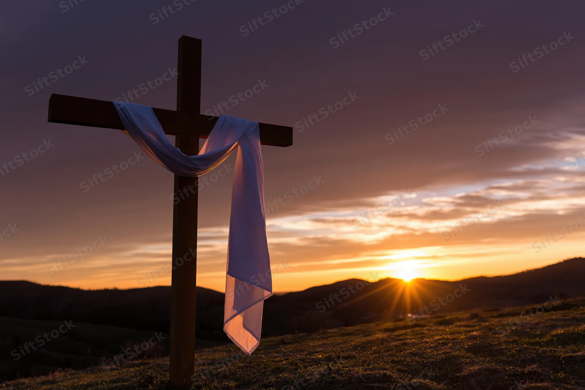 Wooden cross with white cloth draped over it standing on a hill at sunset