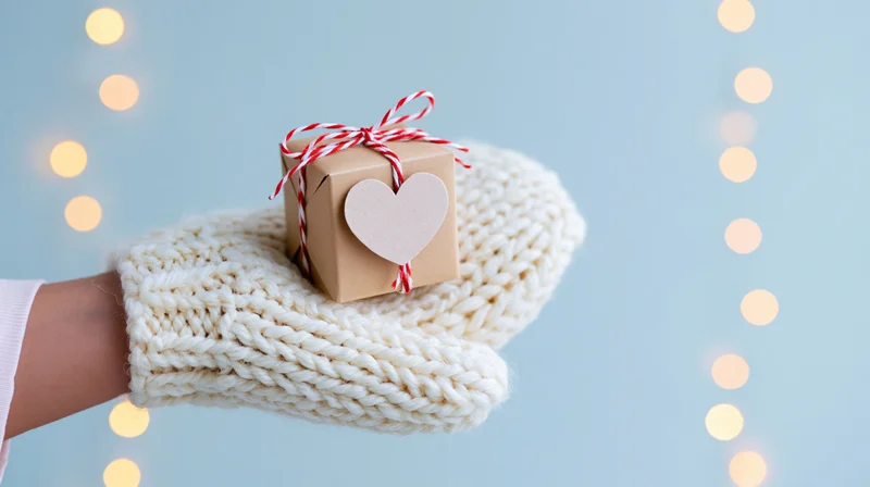 Hand in white knit mitten holding small kraft gift box with heart tag and red striped ribbon against blue background with bokeh lights