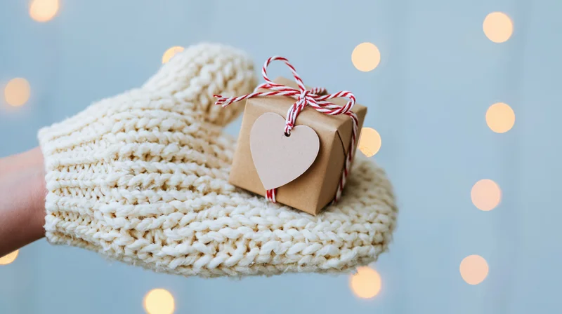 Hand in white knitted mitten holding small kraft gift box with heart tag and red-white string against soft bokeh lights background