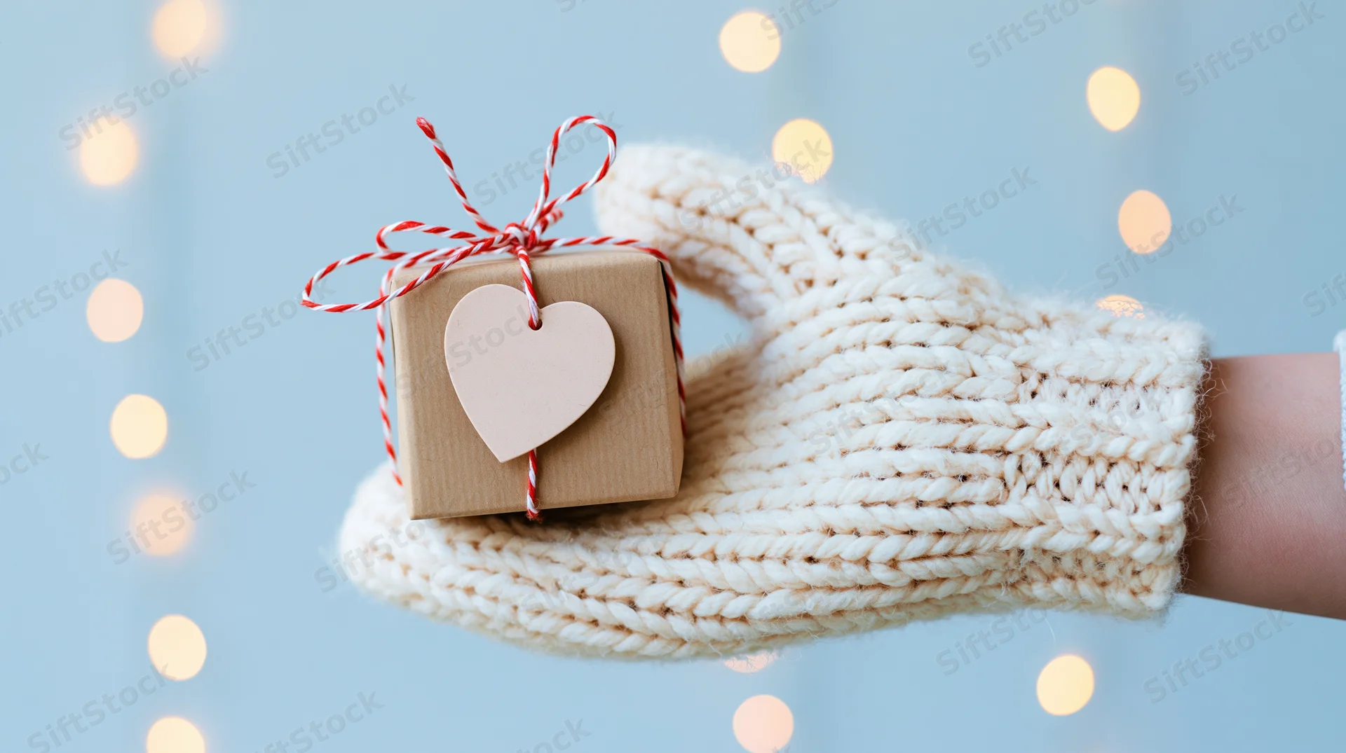 Hand in white knitted mitten holding small kraft gift box with heart tag and red striped string against blue background with bokeh lights
