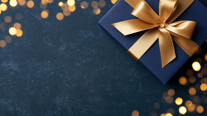 Navy blue gift box with gold satin ribbon bow on dark surface surrounded by warm bokeh lights