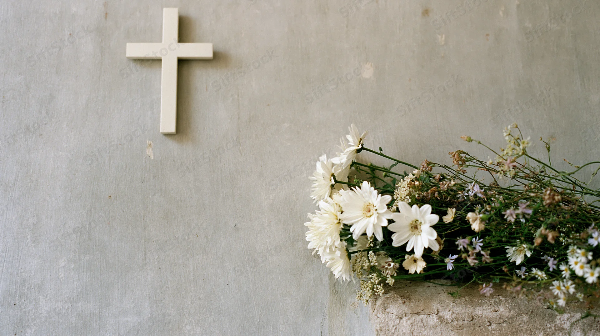 White cross mounted on a gray stone wall beside a bouquet of white daisies and wildflowers