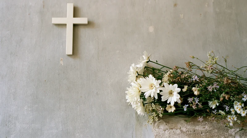 White cross mounted on a gray stone wall beside a bouquet of white daisies and wildflowers