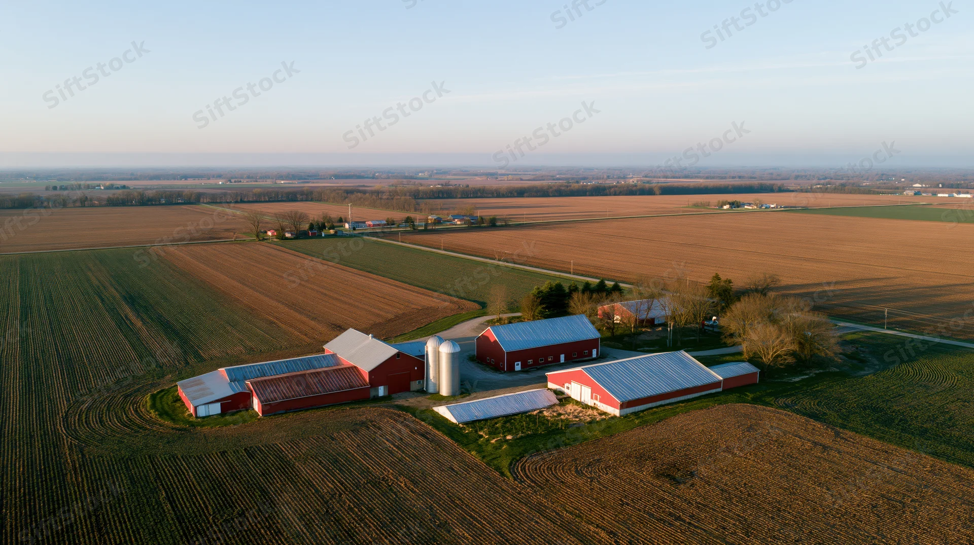 Aerial view of a midwestern farm compound with red barns and silos surrounded by plowed fields at golden hour