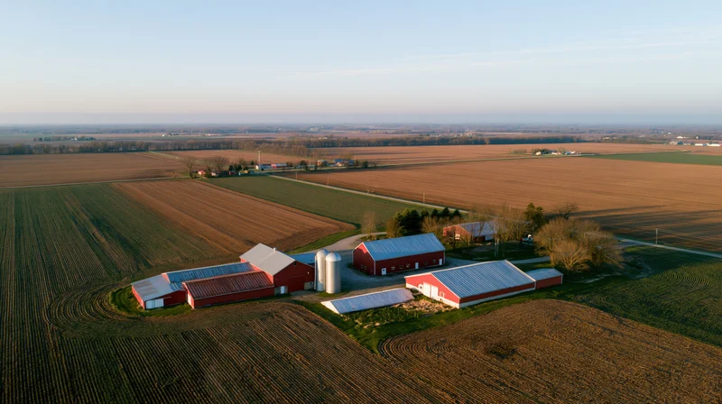Aerial view of a midwestern farm compound with red barns and silos surrounded by plowed fields at golden hour