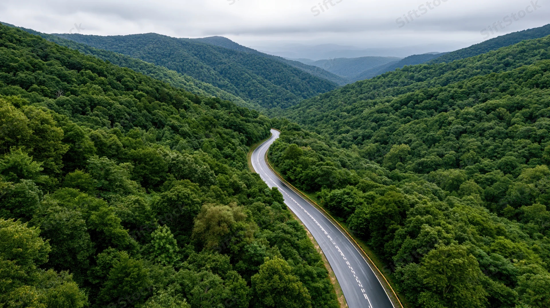 Aerial view of a winding two-lane highway curving through a dense green forest in a mountain valley