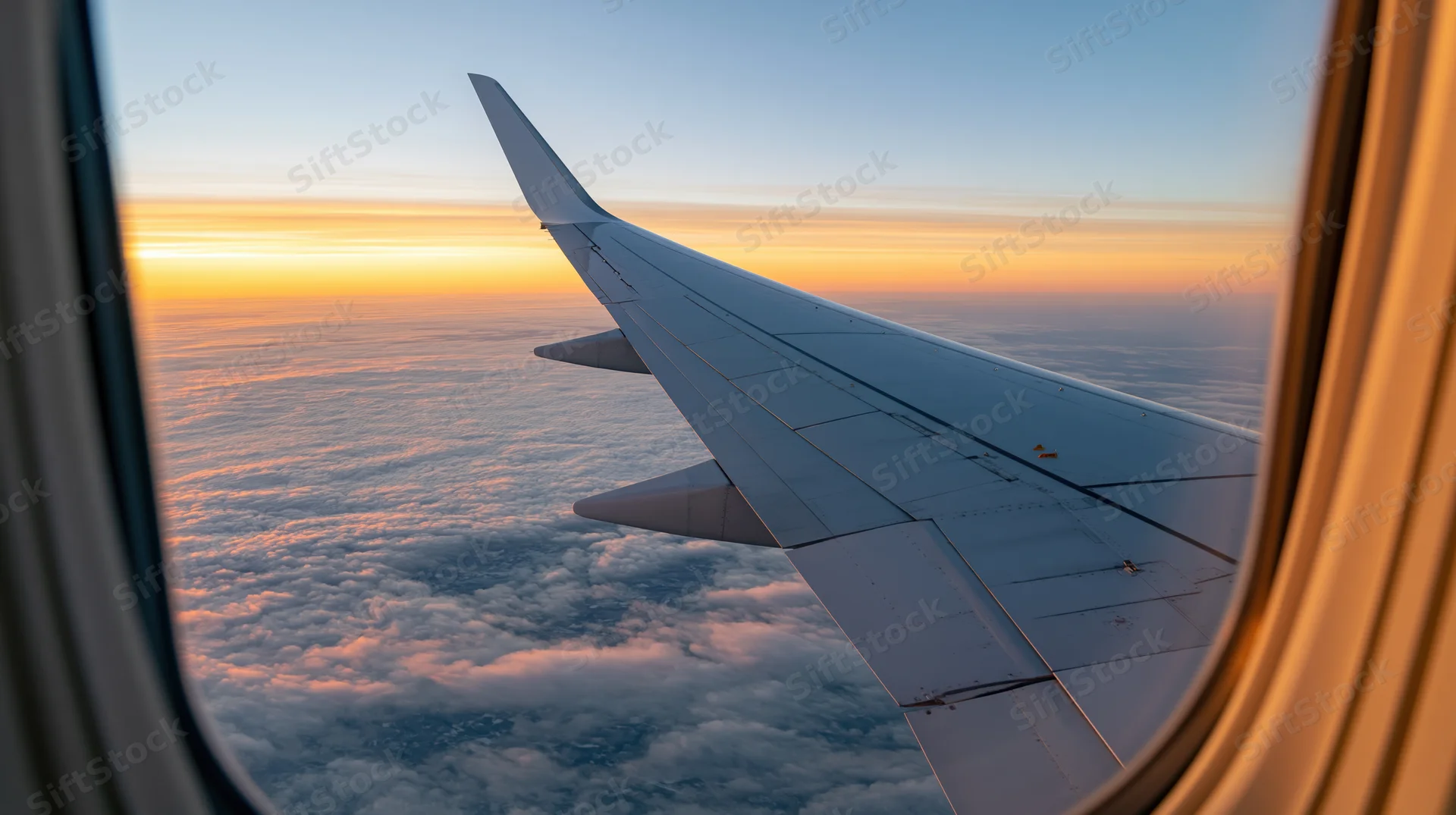 Airplane wing seen through passenger window above clouds at sunset