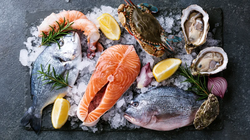 Assorted raw seafood including salmon, oysters, crab, shrimp, and fish arranged on crushed ice with lemon and herbs on a dark slate surface