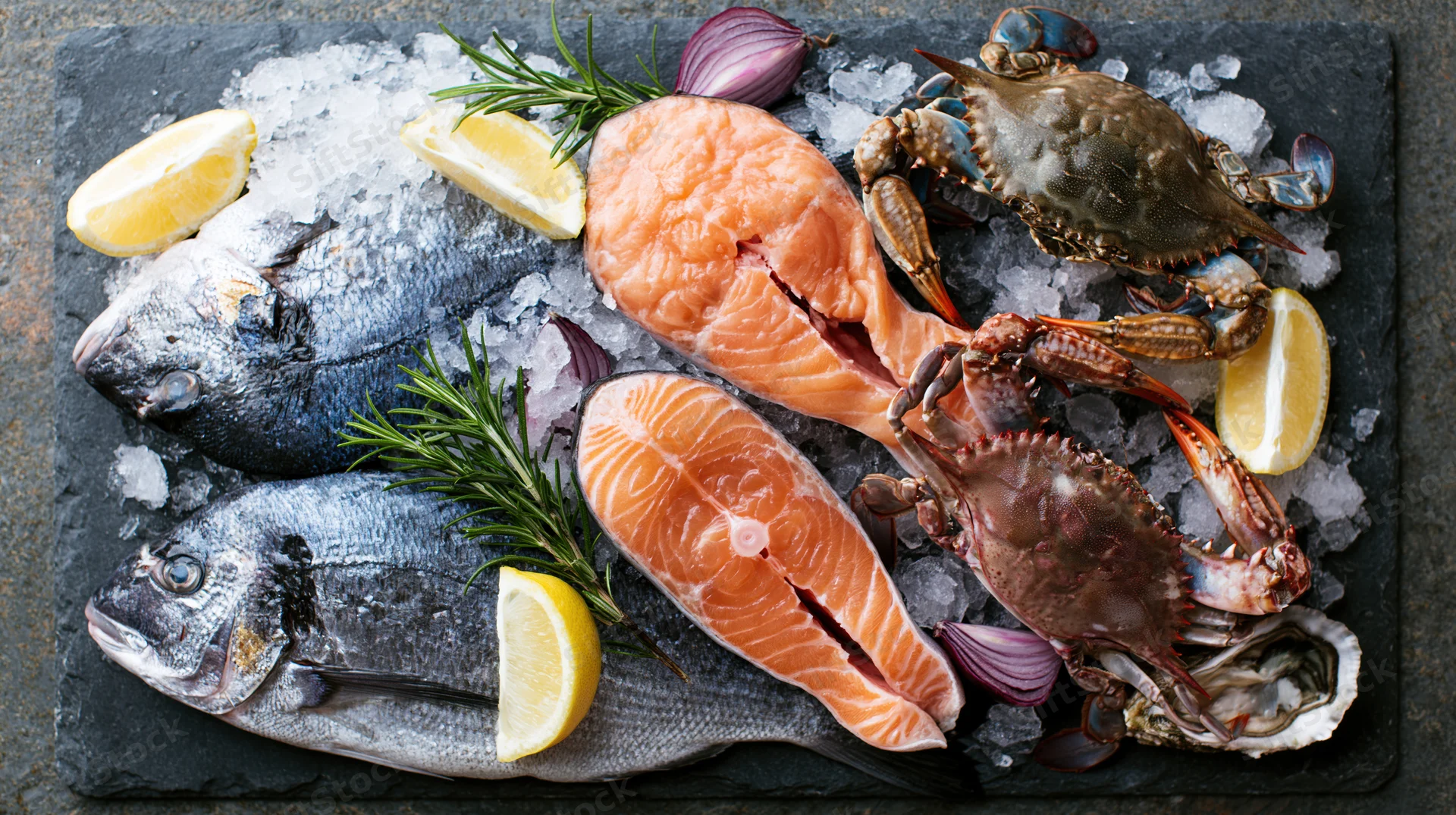 Assorted raw seafood including salmon, crab, oysters and fish arranged on crushed ice on a slate board with lemon and rosemary