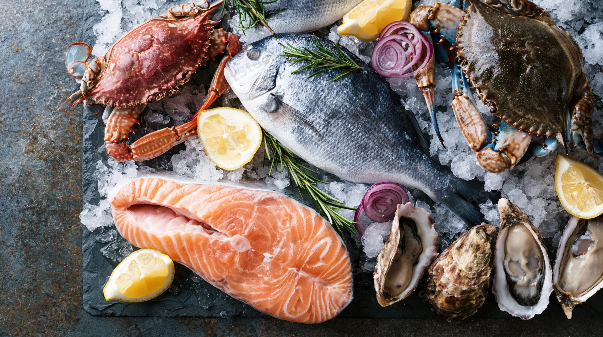 Assorted raw seafood including salmon, crab, oysters, and fish arranged on crushed ice with lemon and rosemary on a dark slate surface viewed from above