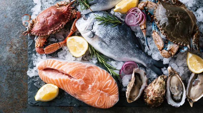 Assorted raw seafood including salmon, crab, oysters, and fish arranged on crushed ice with lemon and rosemary on a dark slate surface viewed from above