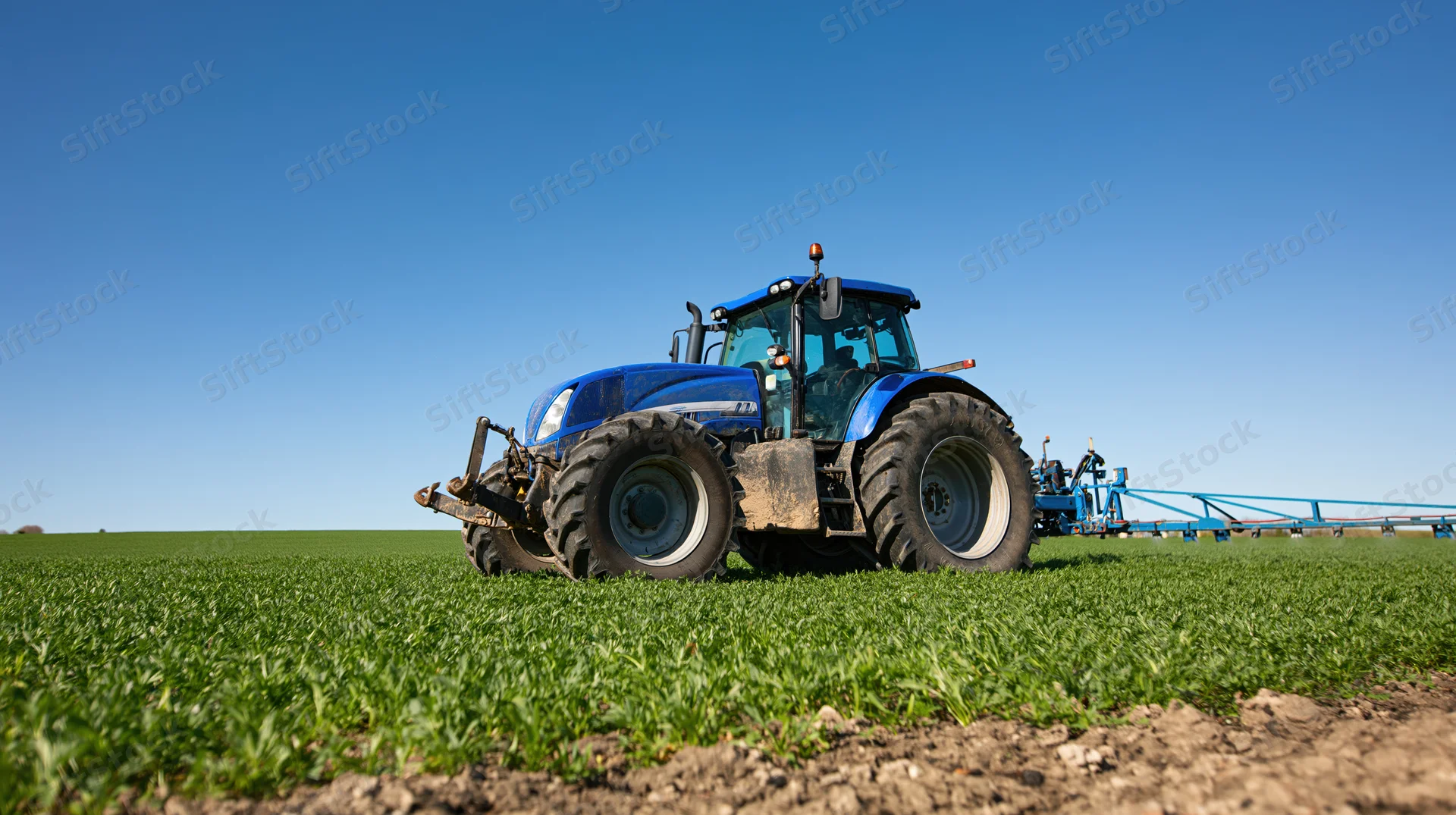 Blue tractor pulling spray boom across green field under clear sky