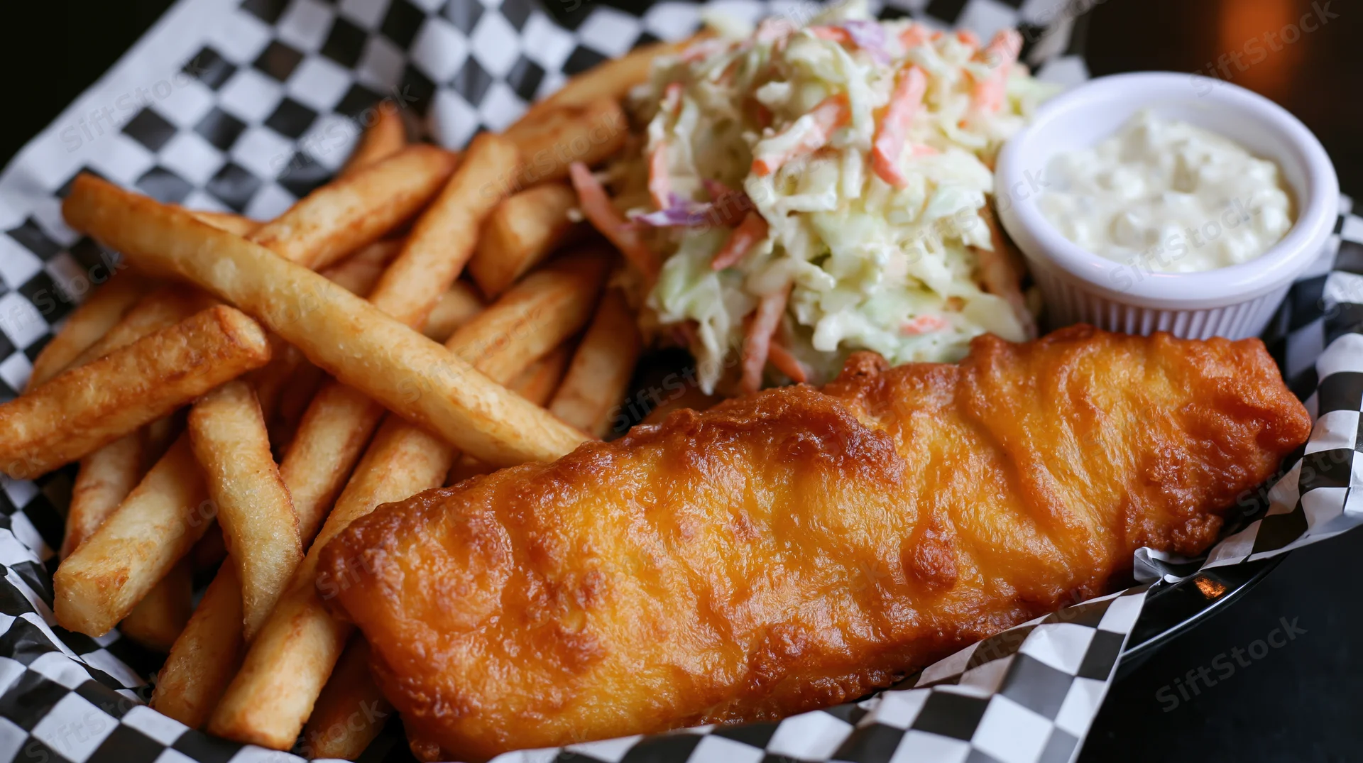 Golden-battered fish and crispy fries served in a basket lined with black-and-white checkered paper with coleslaw and tartar sauce on the side