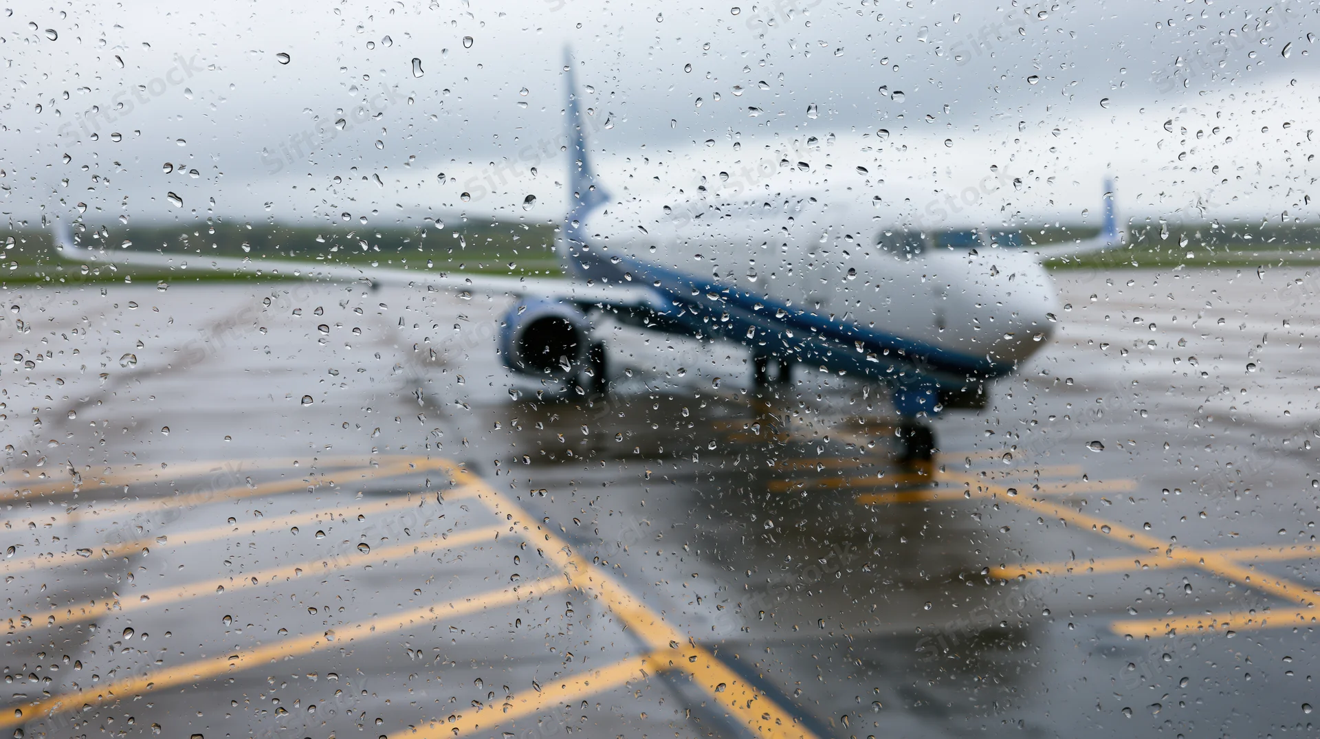 Commercial airplane parked on wet tarmac viewed through rain-covered window