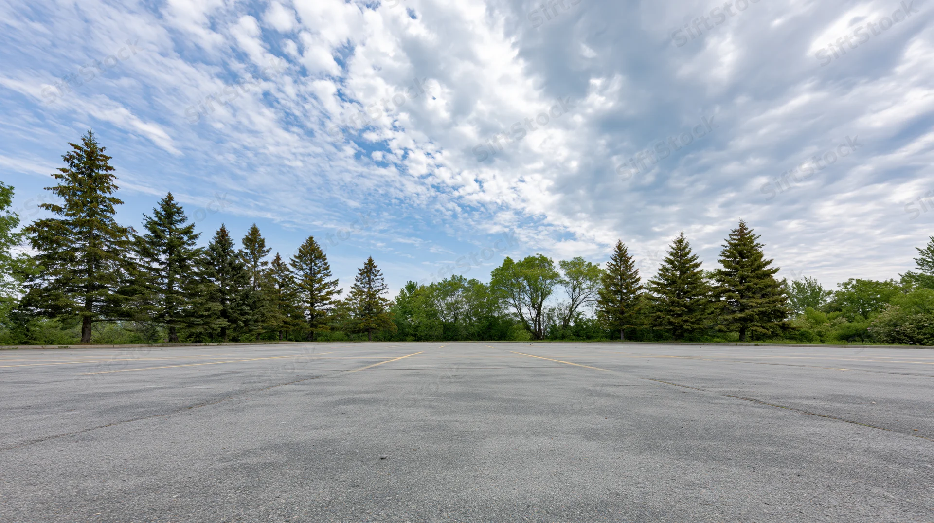Empty asphalt parking lot stretching toward a line of evergreen trees under a partly cloudy sky