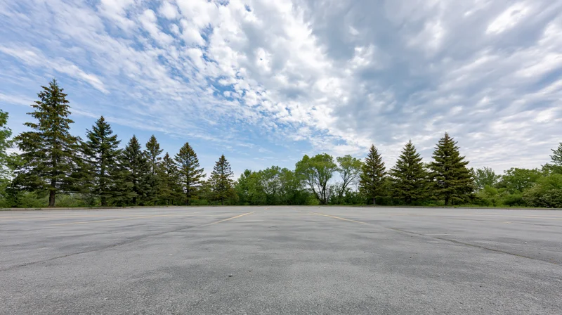 Empty asphalt parking lot stretching toward a line of evergreen trees under a partly cloudy sky