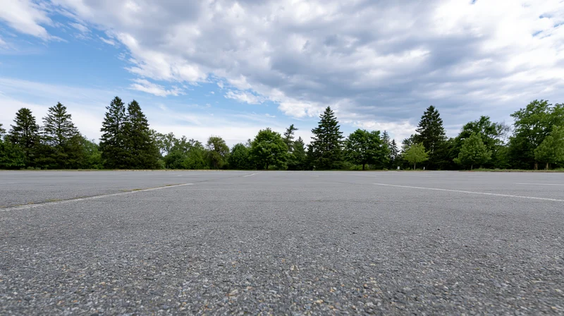 Empty asphalt parking lot stretching toward a line of trees under a partly cloudy sky