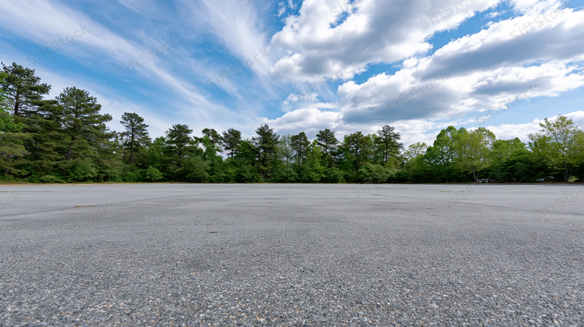 Empty asphalt parking lot stretching toward a line of green trees under a blue sky with white clouds