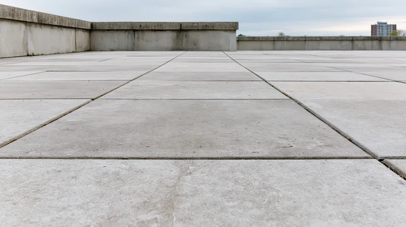 Large square concrete pavers forming an empty urban plaza floor with low walls and distant buildings under an overcast sky