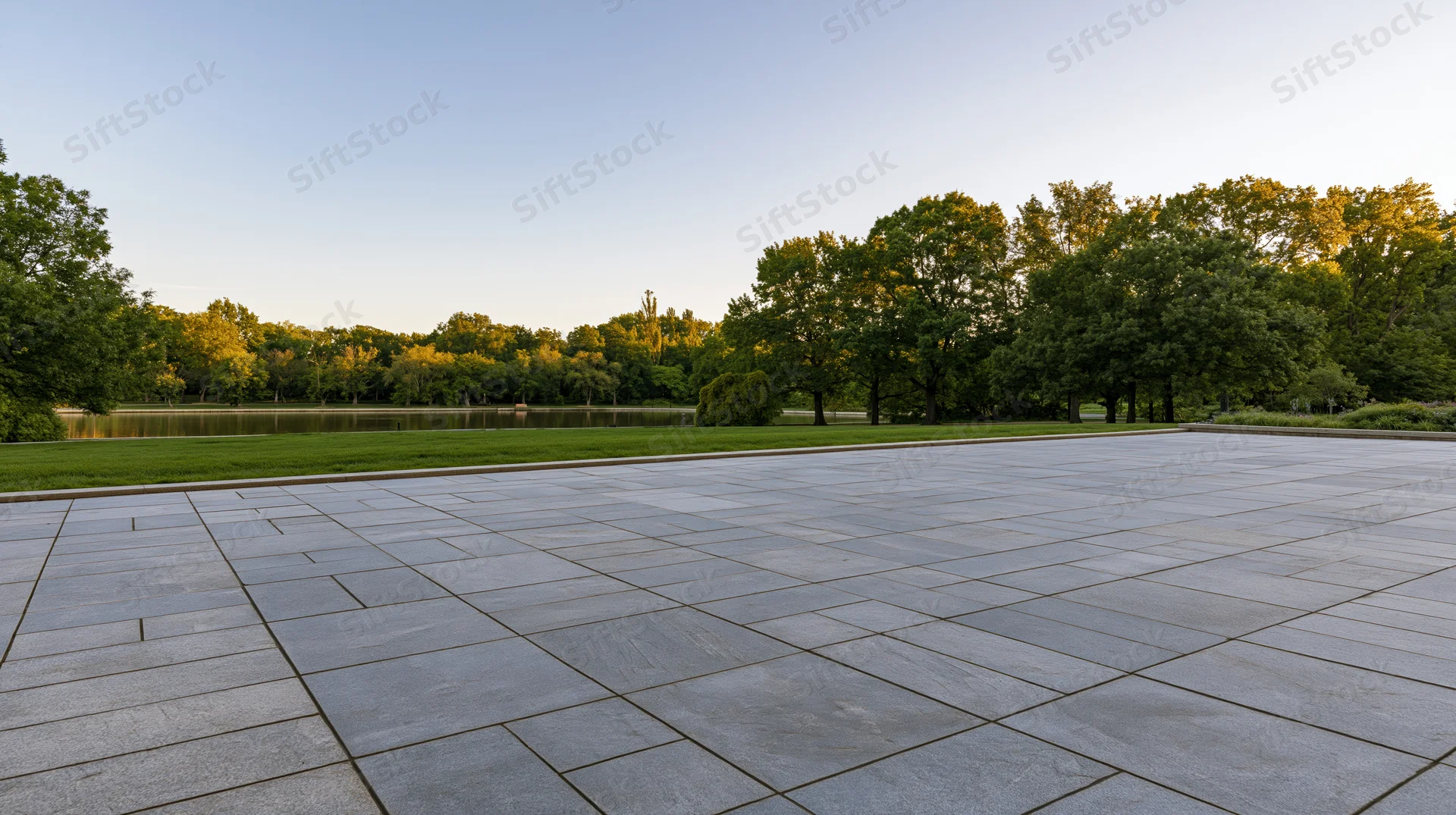 Grey stone tile plaza extending toward a park with trees, grass and pond at sunset