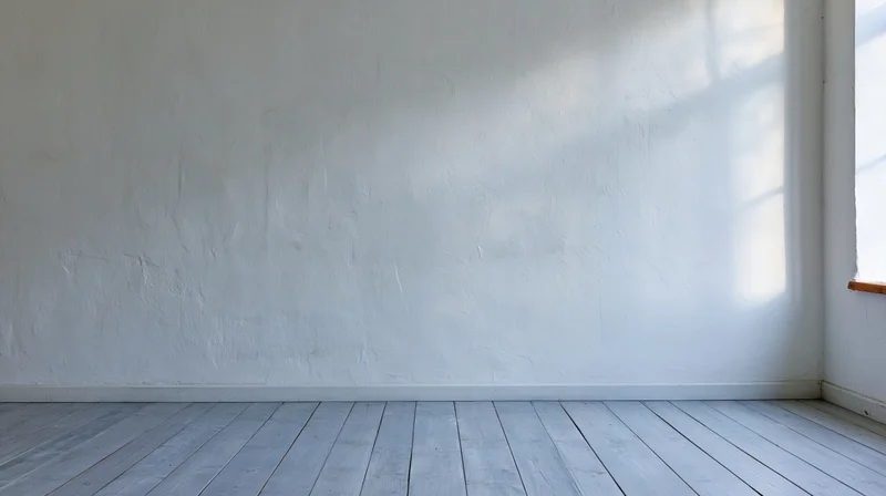 Empty room with white plaster wall and wooden floor lit by natural light from window
