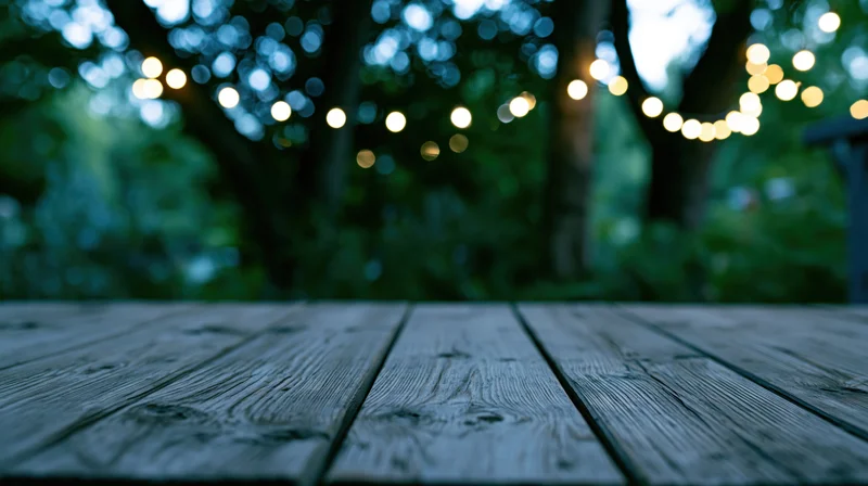 Weathered wooden deck surface in foreground with blurred string lights and trees in background at twilight