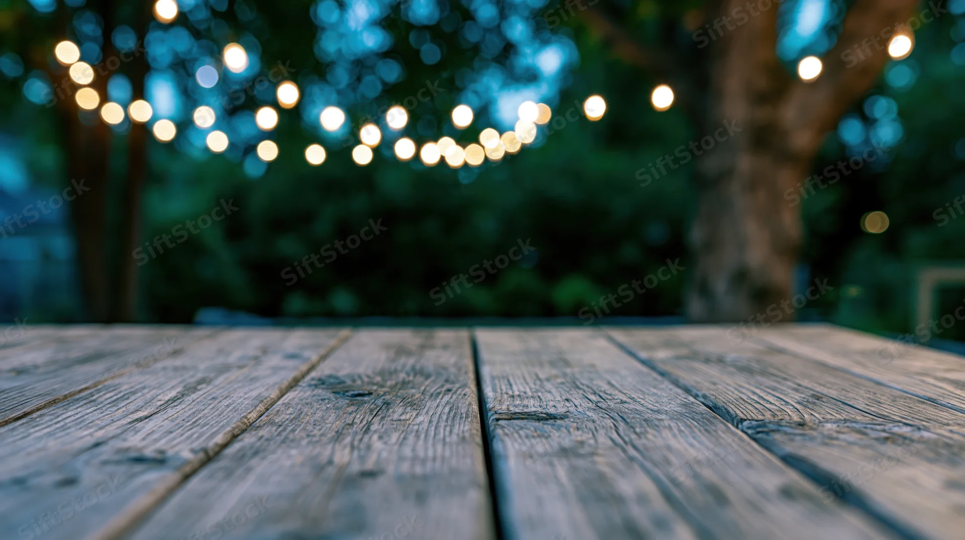 Weathered wooden deck surface in foreground with blurred string lights and trees in background at twilight