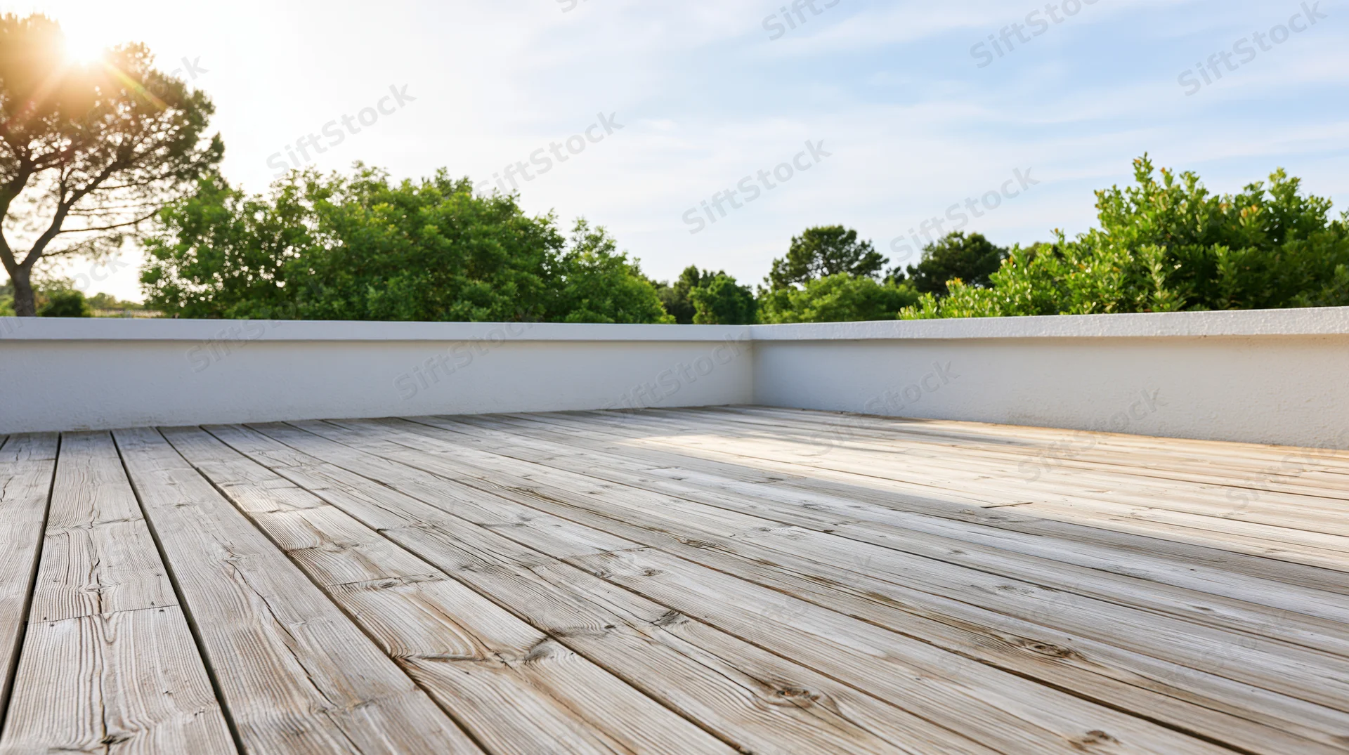 Empty wooden deck terrace with bleached planks and white walls under sunlight, surrounded by green trees and blue sky