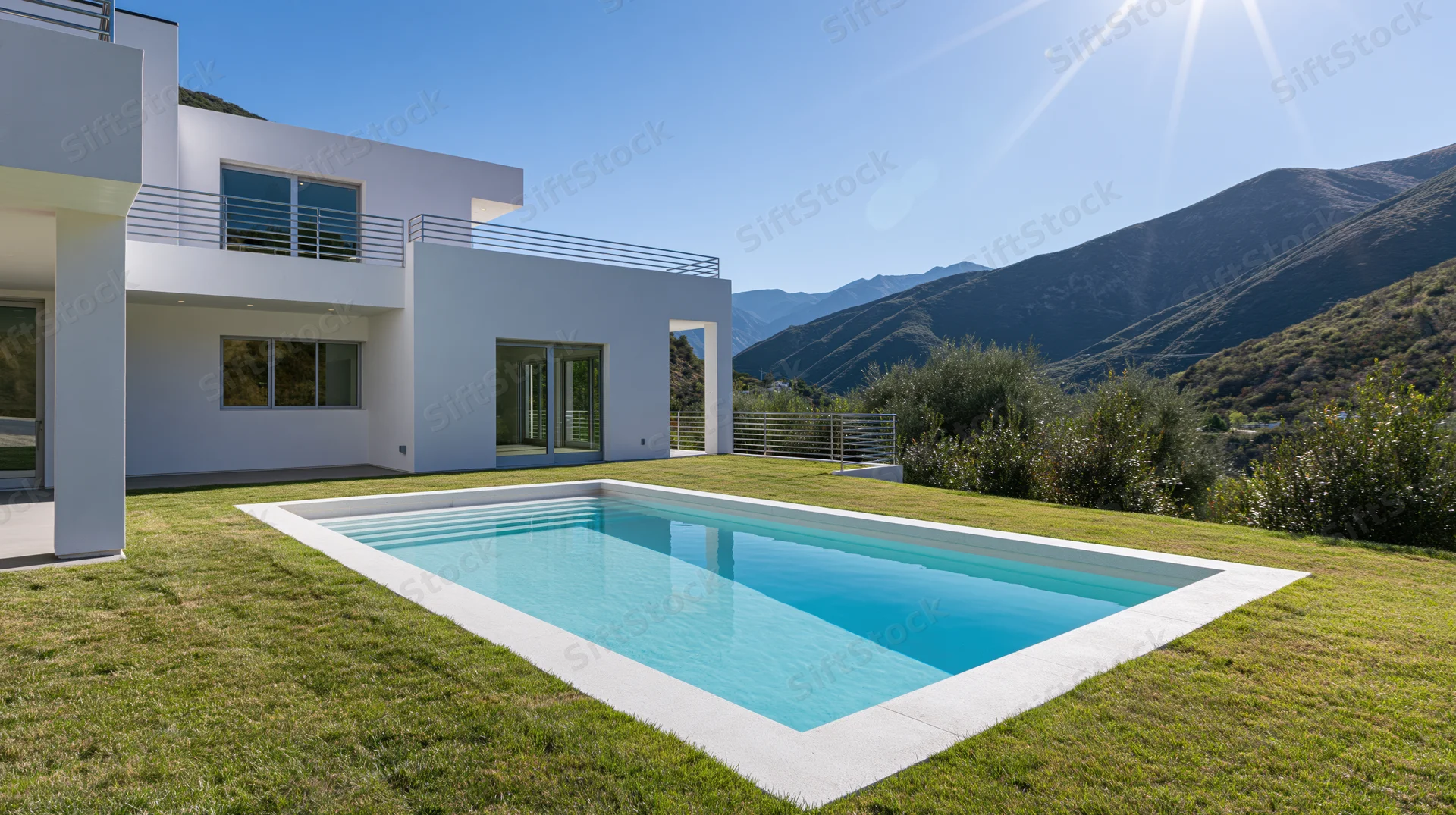 Modern white stucco villa exterior with rectangular swimming pool surrounded by grass and mountains under clear blue sky
