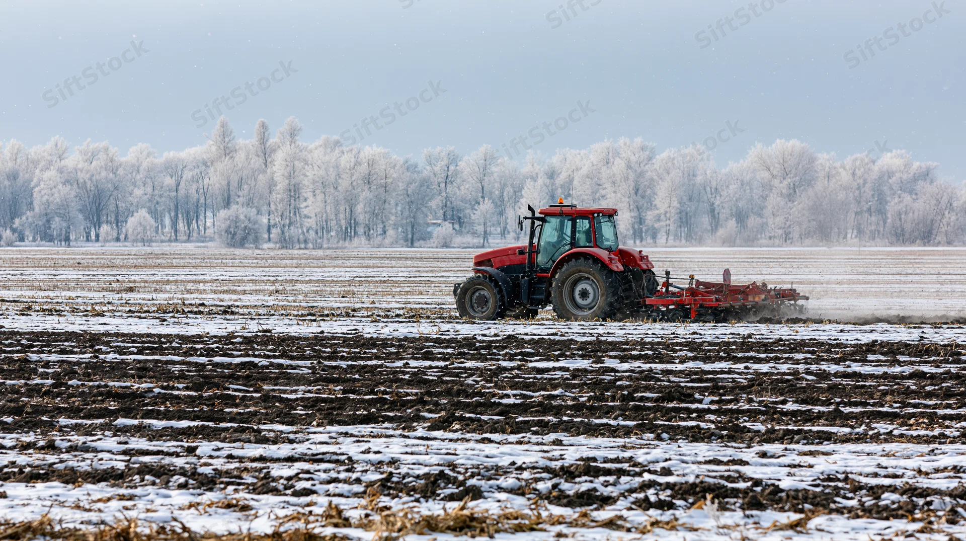 Red tractor pulling tillage implement across snow-dusted field in winter
