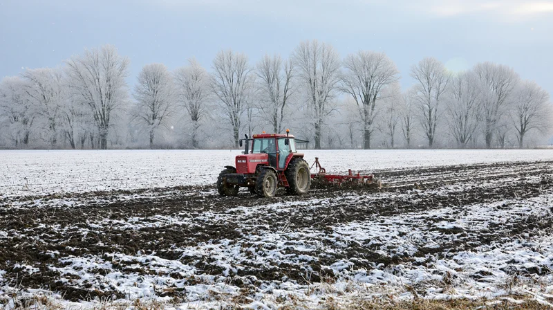 Red tractor pulling tillage implement across snow-covered field in winter