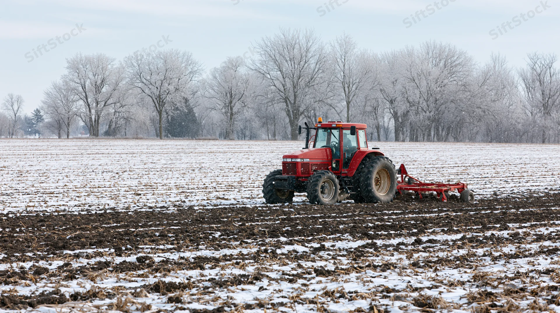 Red tractor pulling tillage implement across snow-covered field in winter