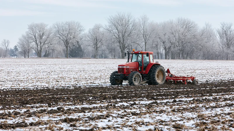 Red tractor pulling tillage implement across snow-covered field in winter
