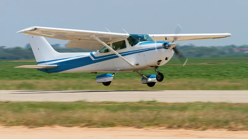 Small white and blue single-engine aircraft landing on a runway with grass field in background