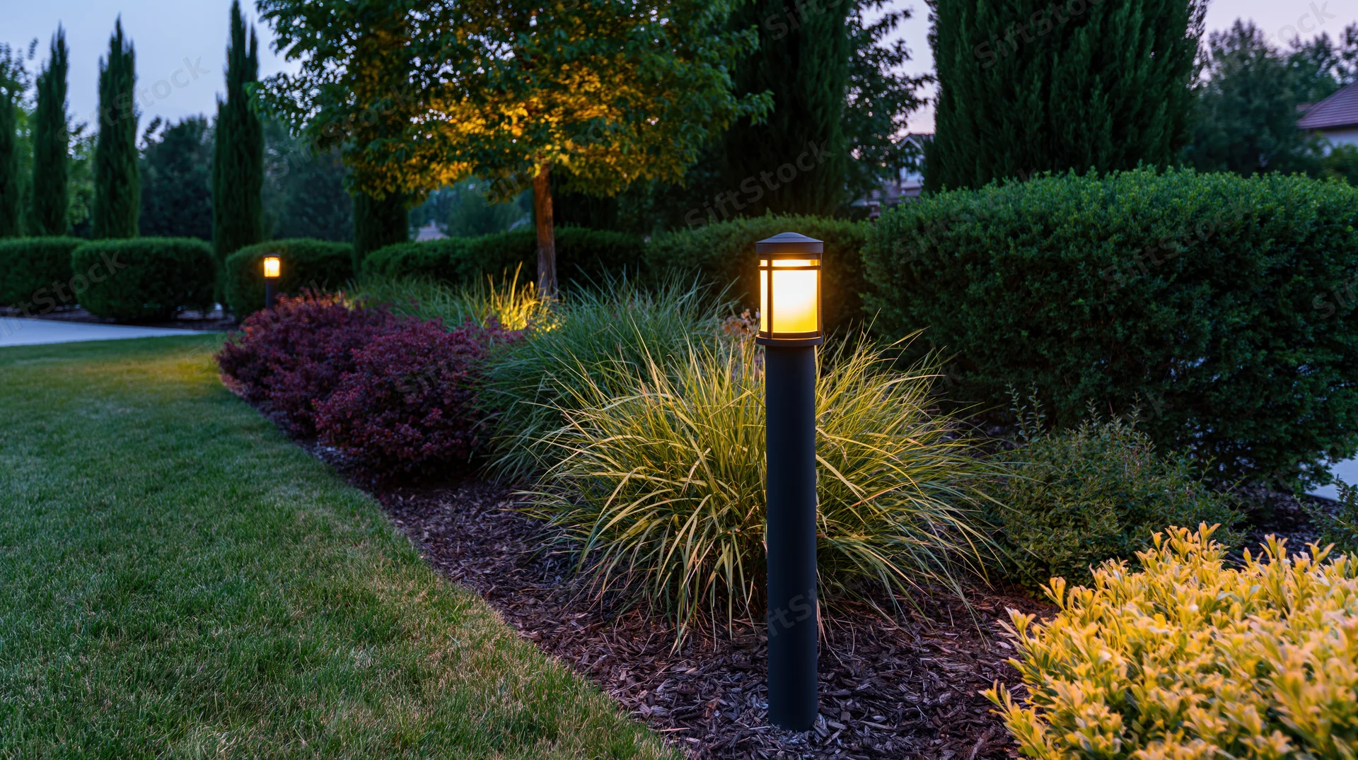 Tall black bollard lights glowing warm amber along a landscaped garden pathway at dusk