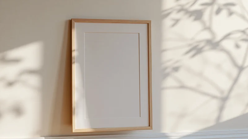 Thin light wood picture frame with white mat leaning against a wall in a sunlit room with leaf shadows