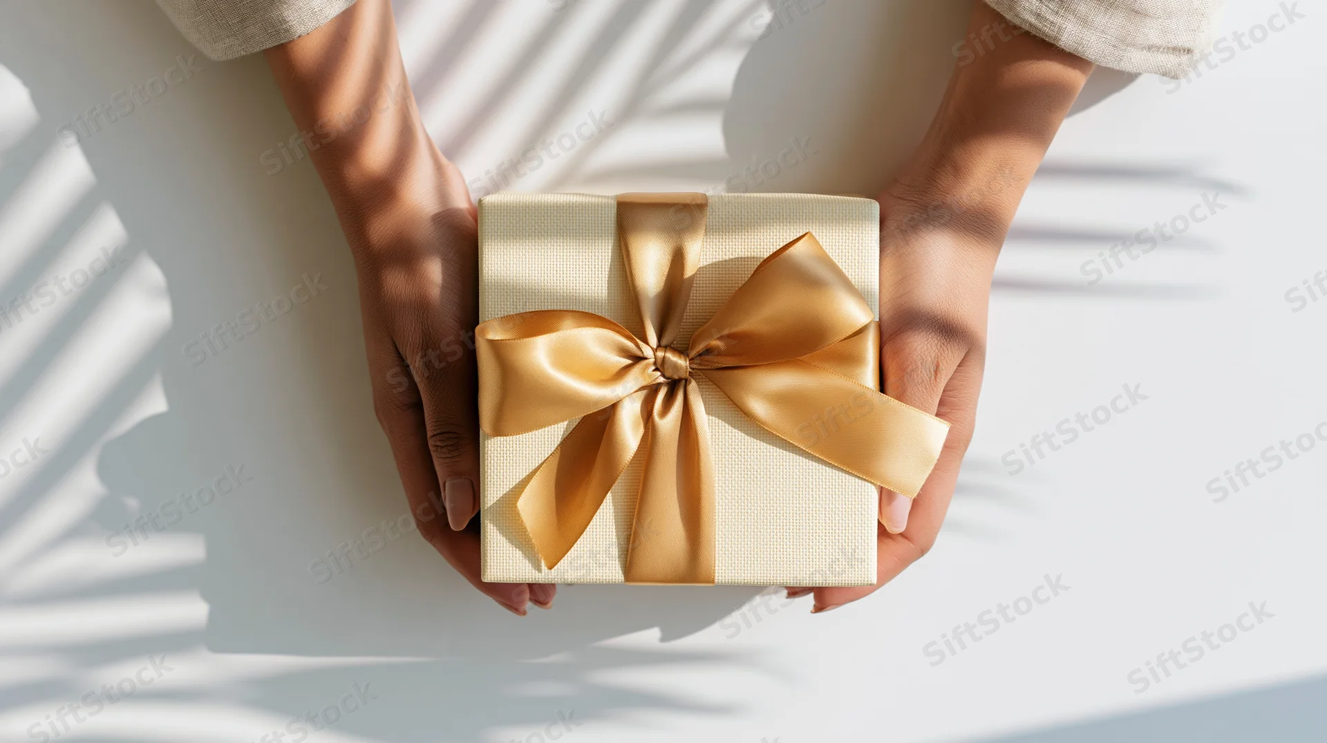 Hands holding a cream gift box with gold ribbon on a white surface with leaf shadows