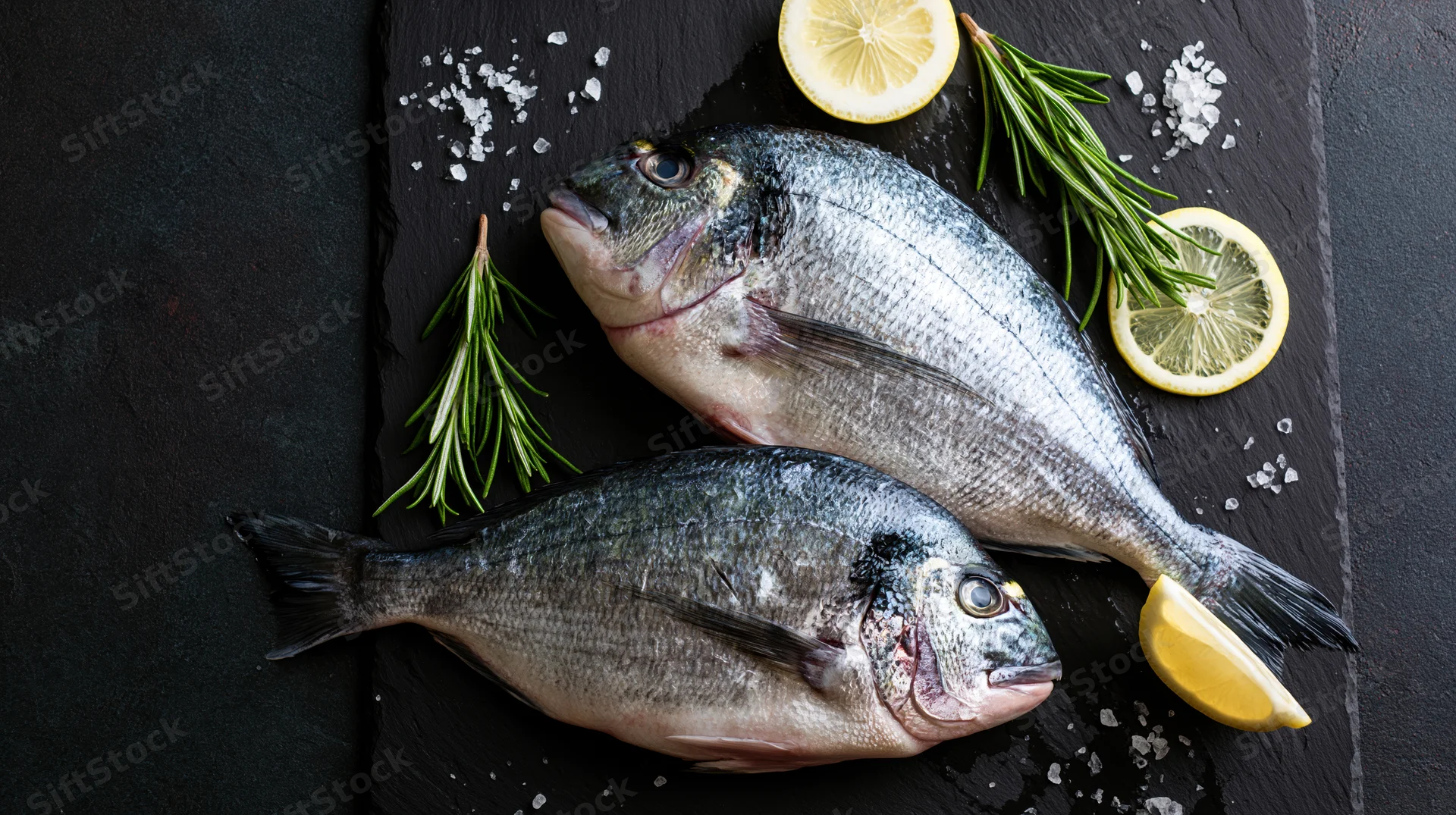 Two raw whole sea bream fish on a dark slate board with lemon slices, rosemary sprigs and coarse salt, arranged for culinary presentation
