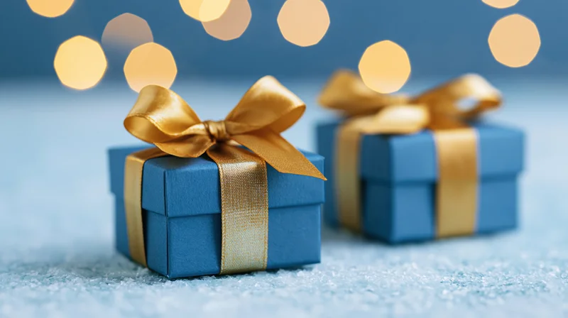 Two blue gift boxes with gold satin ribbon bows on a snowy surface under warm bokeh lights