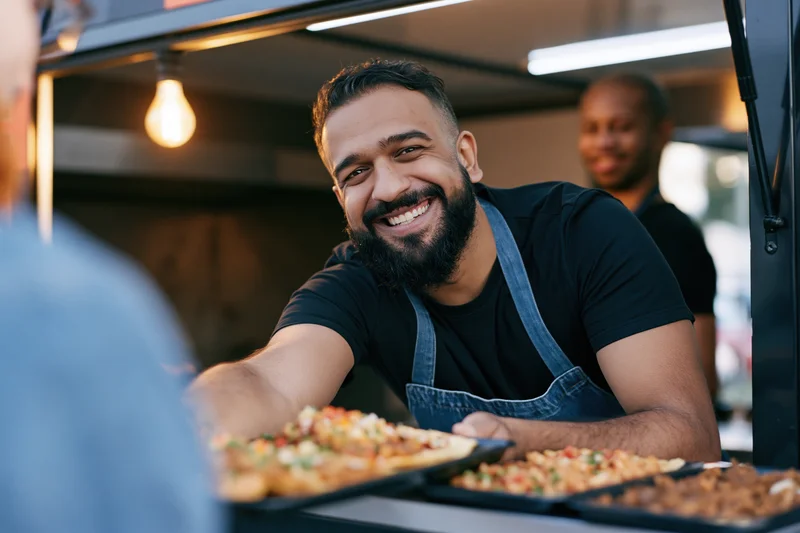 Bearded man in apron smiling while serving food from a food truck window