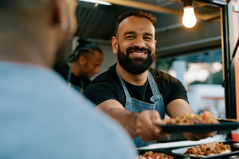 Bearded man in denim apron serving food from food truck window to customer