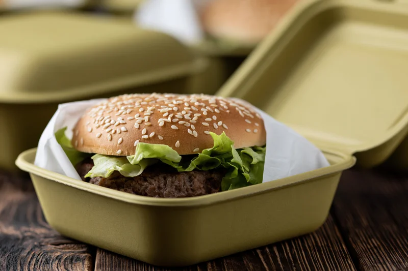 Beef burger with sesame seed bun and lettuce in a green takeout container on wooden table
