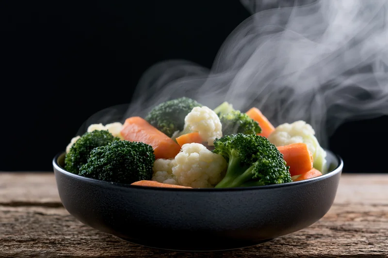 Steamed broccoli carrots and cauliflower in a black ceramic bowl on a wooden table
