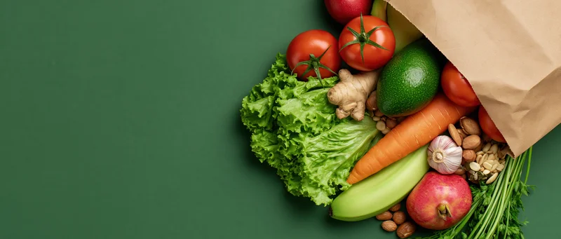 Brown paper grocery bag tipped over spilling fresh vegetables and fruits on green surface