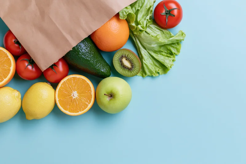 Brown paper bag spilling fresh fruits and vegetables on blue surface