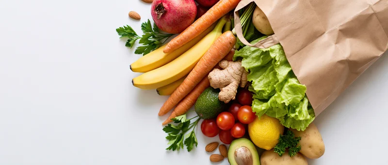 Brown paper grocery bag tipped over spilling fresh fruits and vegetables on white surface