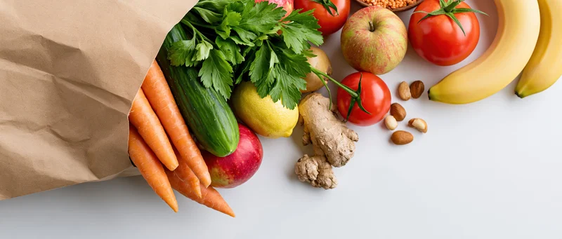 Brown paper bag spilling fresh vegetables and fruits on white surface
